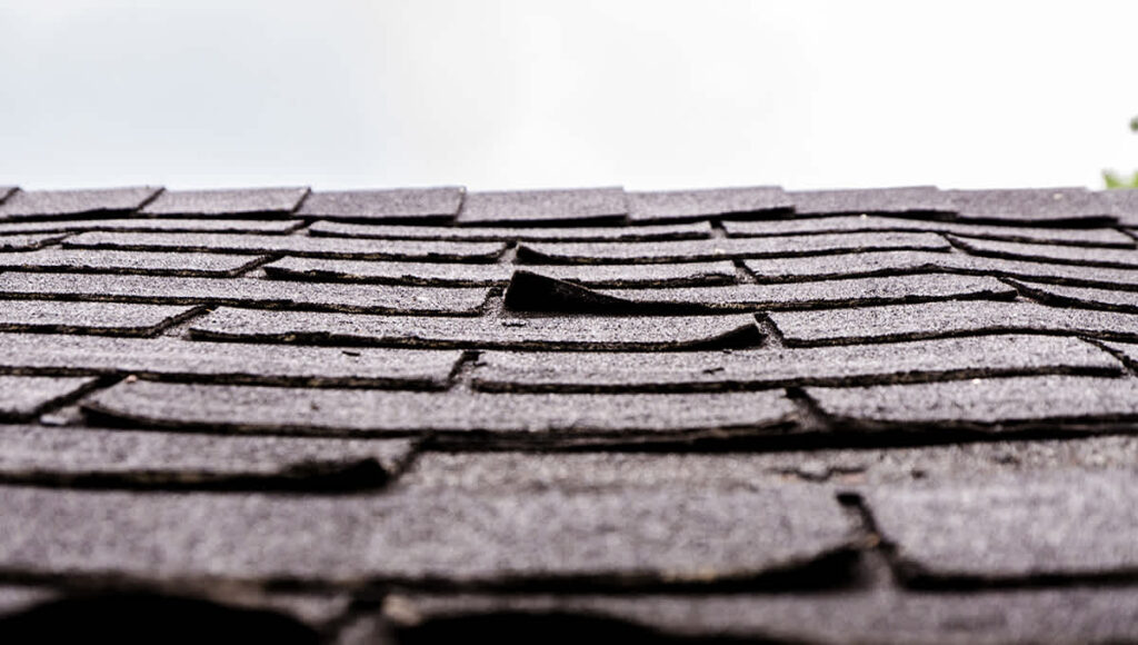 Close up up dark grey shingles that are cupping on the corners of the shingle tabs. A roof repair or roof replacement is needed.